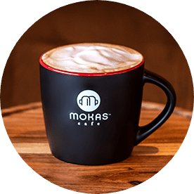 A black mug with the Mokas logo on the front, filled with coffee, topped with foam.
