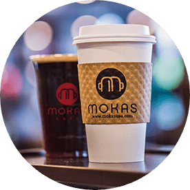 Two coffee cups, one iced dark cold brew without a lid in a plastic cup with the Mokas logo on the front and one hot to-go white coffee cup with the Mokas logo on the brown coffee sleeve.