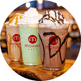 Three different frappé, one in front of the other. Starting from the furthest, a caramel frappé with whipped cream and caramel drizzle, a green frappé with whipped cream and a chocolate frappé with whipped cream and chocolate drizzle.