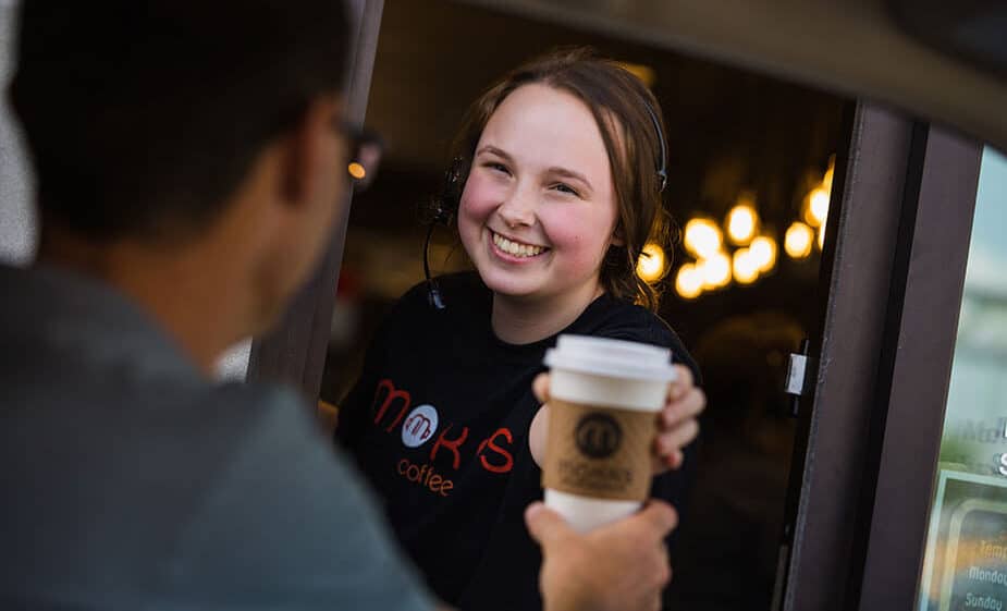 A Mokas employee smiling handing a man his hot to-go coffee from the drive-thru window.